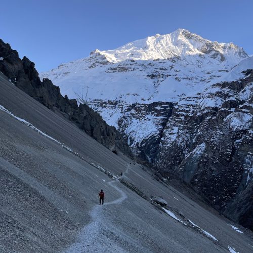 A person hiking along a narrow path in a snowy mountain landscape, under a clear sky.