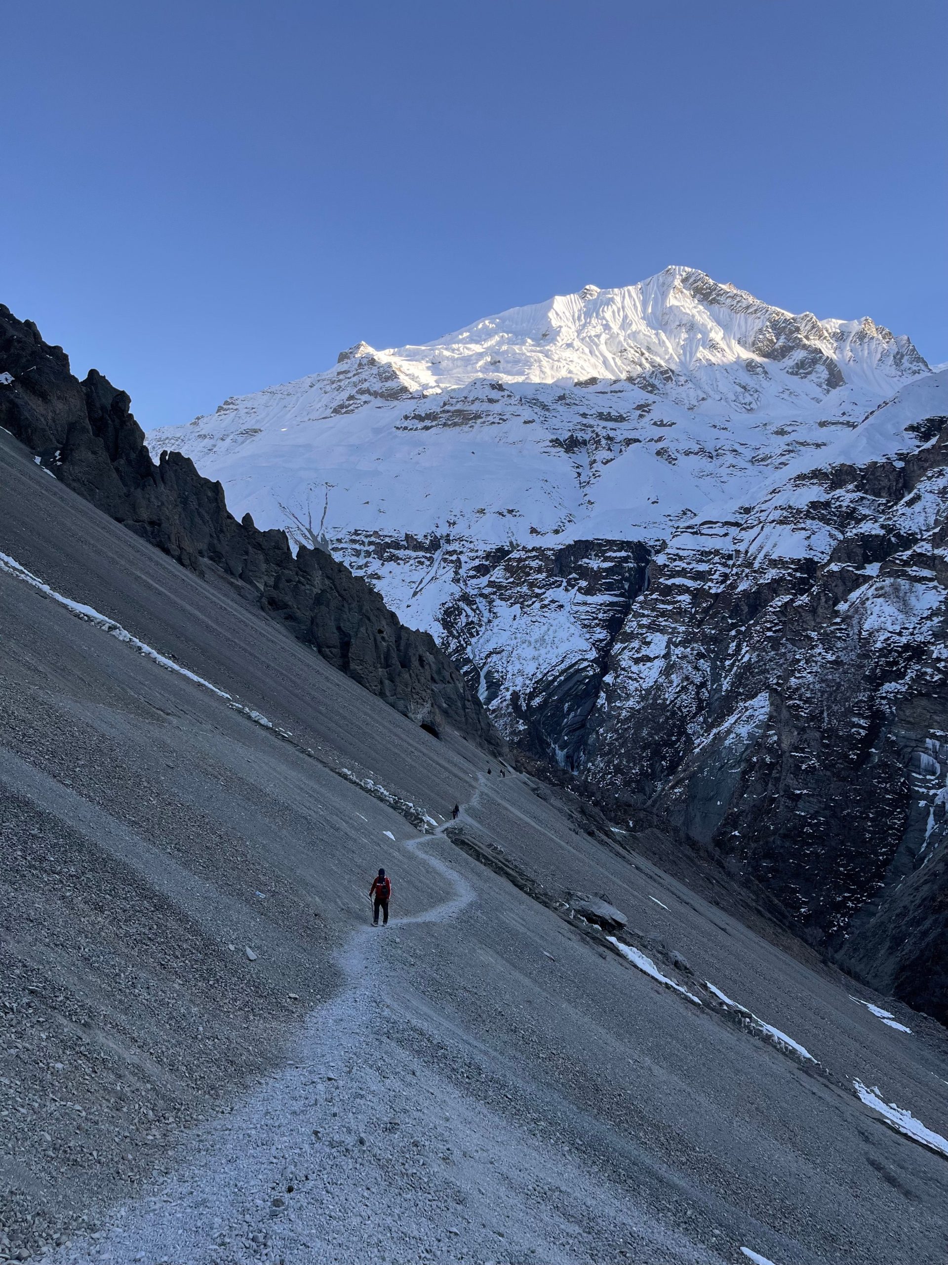 A person hiking along a narrow path in a snowy mountain landscape, under a clear sky.